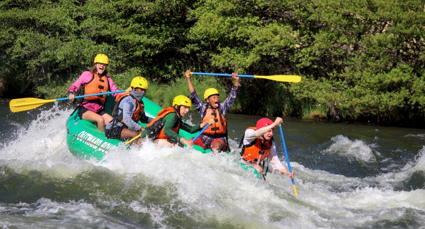 A group of students wearing safety gear paddle a raft through whitewater. One of them lifts their paddle into the air in apparent joy.